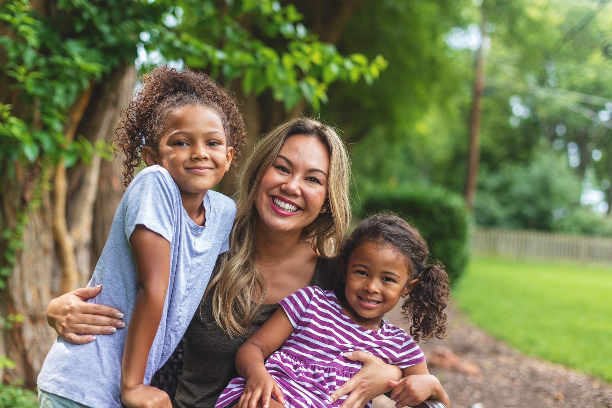Mother with two daughters of mixed Chinese and African American ethnicity in a green lush back yard setting posing for portraits smiling and being silly