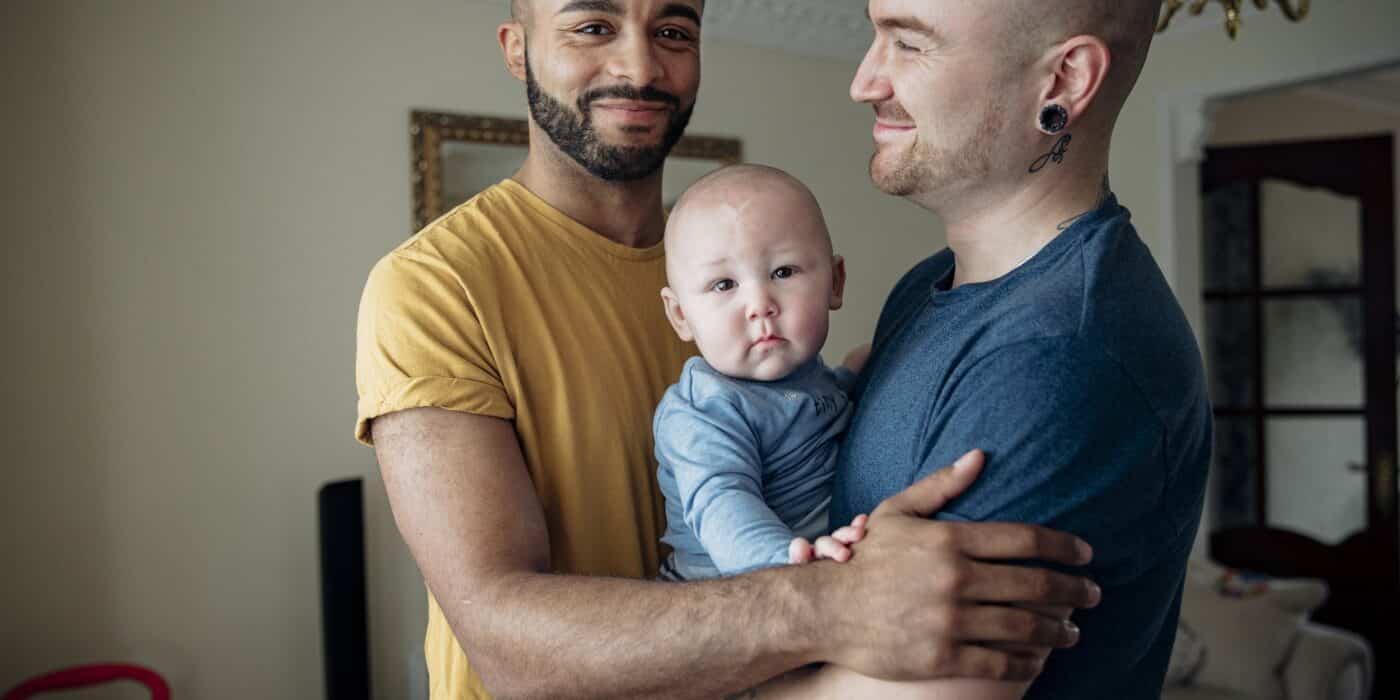 Two men spending time with their little baby boy. They're all together at home standing in an embrace