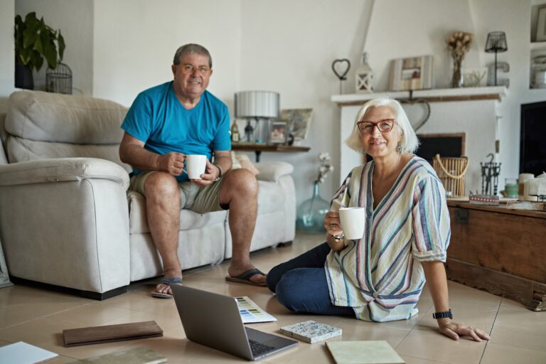 Couple in 50s and 60s wearing casual clothing and sitting in family living room with color swatches, tile samples, and coffee to inspire ideas for home improvement.
