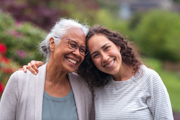 A senior woman embraces her millennial Eurasian daughter as they happily walk through a natural parkland area and enjoy their time together. The daughter is smiling at the camera.